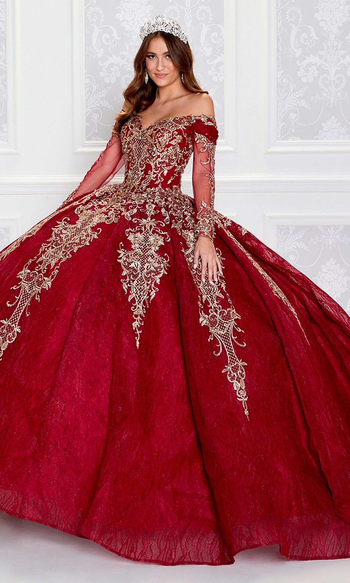 A woman wearing the Princesa Off-the-Shoulder Lace Quince Dress PR12267 with gold embroidery, a tiara, and a lace-up corset back poses in front of a white patterned wall.